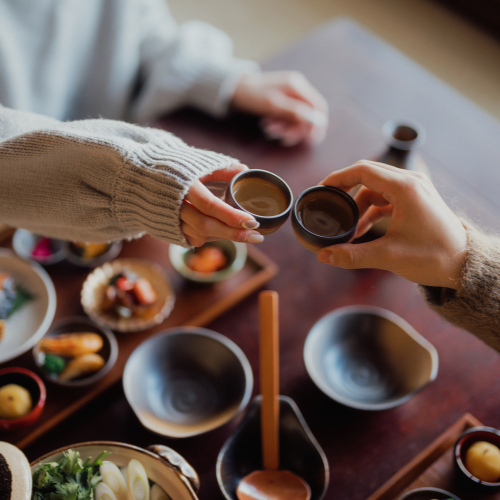 Two people clinking sake cups over a table filled with assorted Japanese dishes in a cozy dining setting