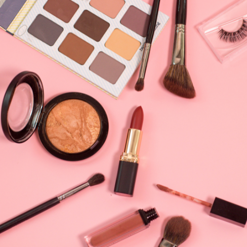 Flat lay of makeup products including eyeshadow, lipstick, and brushes on pink background