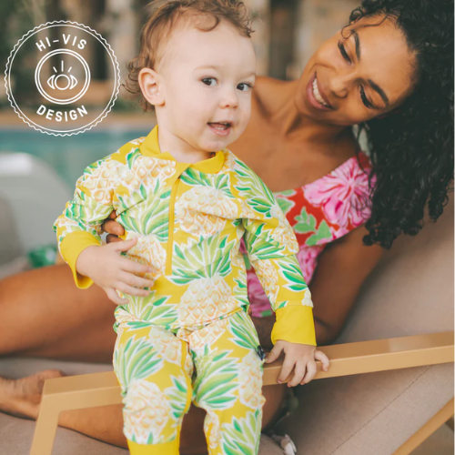 Child wearing a yellow pineapple-patterned Honeysuckle Swim Co. outfit with a woman in a pink swimsuit, both sitting on a deck.