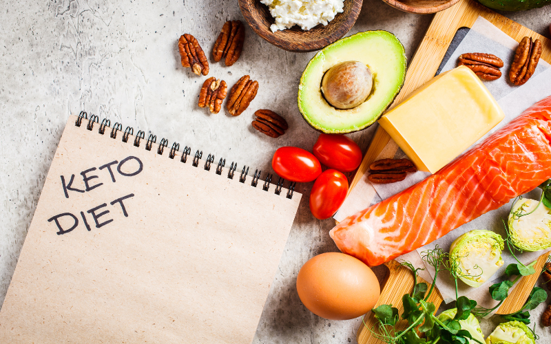 Keto diet food items including salmon, avocado, cheese, and vegetables on a concrete surface.