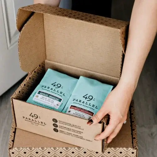Cardboard box with blue packages labeled '49 Parallel' being handled.