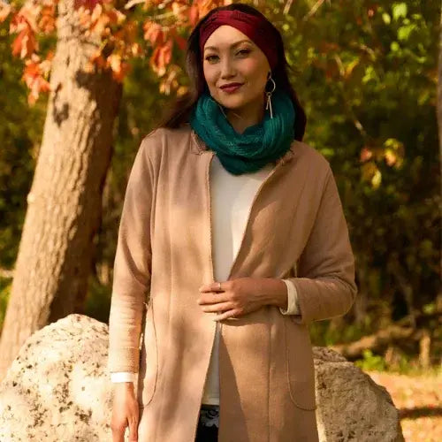 Woman wearing a beige coat and teal scarf standing outdoors with trees in the background