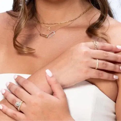 Close-up of a woman wearing layered gold necklaces, including one with an initial 'A' charm and a mix of paperclip and pavé styles. She also wears several gold rings with diamonds on her fingers, along with a strapless white top and a soft pink manicure
