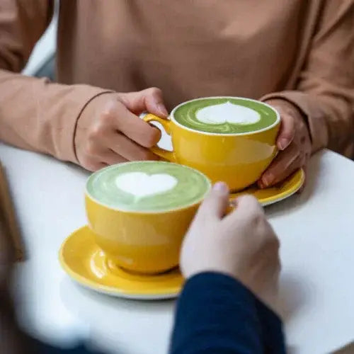 Two people holding yellow mugs with green drinks on a white table.