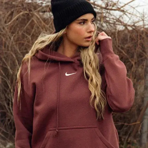 A woman wearing a black beanie and a maroon Nike hoodie stands outdoors in front of leafless branches. 