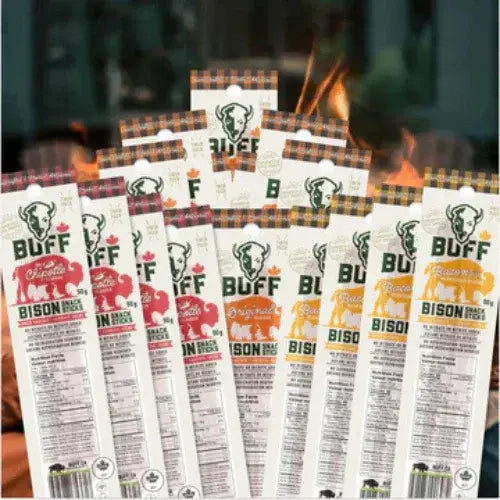 A variety of BUFF brand bison snack sticks displayed in front of a campfire. The packaging features plaid accents and different flavour labels, including Chipotle, Original, and Roasted Garlic. Each package highlights nutritional information and a Canadian maple leaf icon.