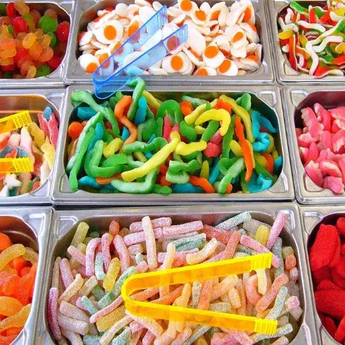 Assorted colourful gummy candies in metal trays with yellow tongs.