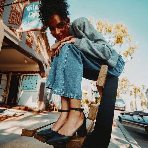 Woman wearing black Call It Spring heels, blue jeans, and a grey sweater sitting on a modern wooden bench outside on a sunny day