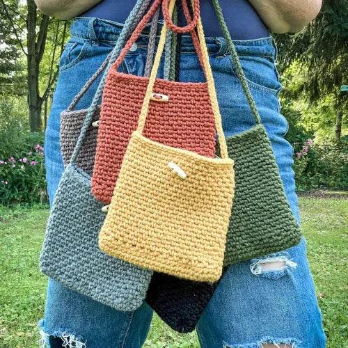 Person holding multiple crocheted bags of different colors in a garden setting