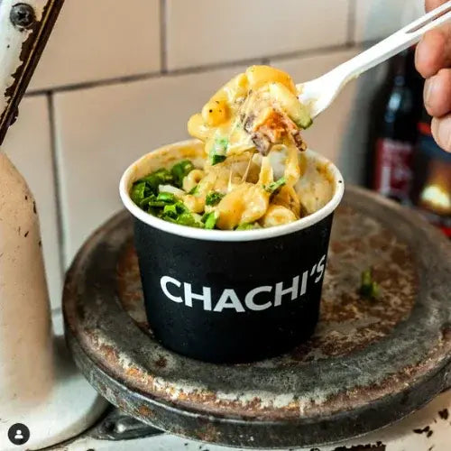 A black takeout cup labeled 'CHACHI'S' filled with creamy macaroni and cheese, garnished with chopped green onions and chunks of meat. A hand lifts a forkful of the cheesy pasta, with melted cheese stretching from the cup, all placed on a rustic metal scale.
