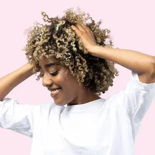 Smiling woman with curly highlighted hair wearing a white t-shirt, holding her hair with both hands against a soft pink background.