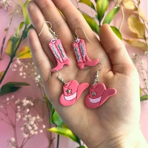 Pink cowboy boot and hat earrings held in a hand with a floral background