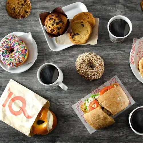 Flat lay of a breakfast spread on a dark wooden table, featuring coffee cups, a sandwich, a pretzel bagel in branded Coffee Time packaging, assorted cookies, a muffin, a granola-covered donut, and a sprinkle-covered donut. The scene captures a variety of sweet and savoury options.