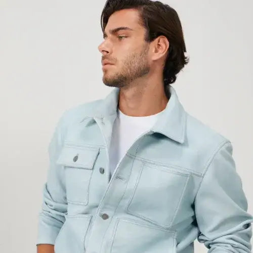 A man with short dark hair and a trimmed beard wearing a light blue utility-style jacket over a white T-shirt. 