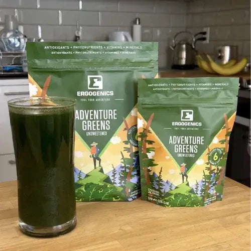 Two packages of Ergogenics Adventure Greens on a kitchen counter with a glass of green juice.