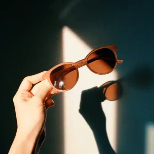 Hand holding a pair of round brown sunglasses casting a shadow on a sunlit wall – Stylish eyewear from a Canadian brand