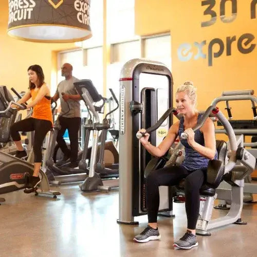 People working out in a bright and modern gym, including a woman using a chest press machine and two others on cardio equipment. The space features yellow walls with 'Express Circuit' signage and various strength and fitness machines