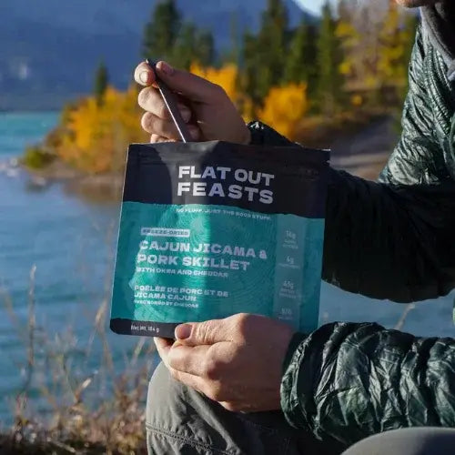 Person holding a Flat Out Feasts food package by a scenic lakeside with mountains in the background.