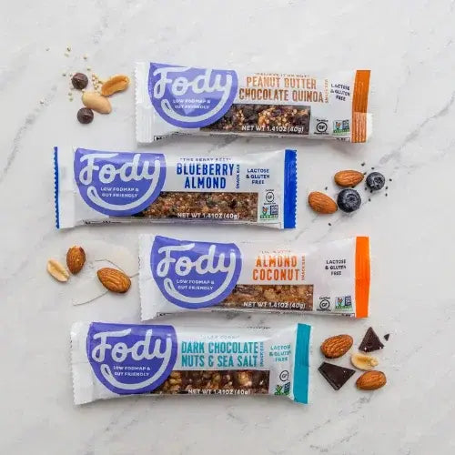 Four Fody brand nut bars on a marble surface with ingredients around them.