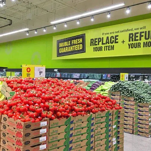 FreshCo produce section featuring a large display of tomatoes and cucumbers under a bright green wall with a 'Double Fresh Guaranteed' sign offering item replacement or refund.