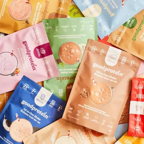 Close-up of assorted GoodProtein all-in-one shake packets in various flavours, laid out flat with colourful packaging and images of chocolate shakes with striped straws.