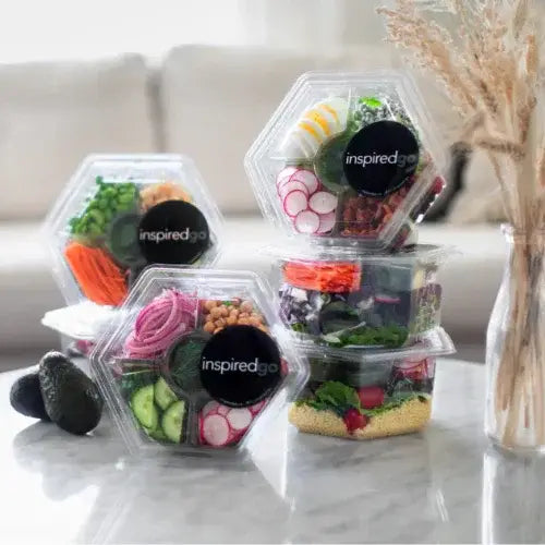 A variety of colourful pre-packaged salads from Inspired Go displayed on a marble table. Each clear hexagonal or round container shows fresh ingredients like radishes, chickpeas, avocado, eggs, greens, and grains. A pair of avocados and a decorative dried plant sit nearby, with a neutral-toned couch in the background.
