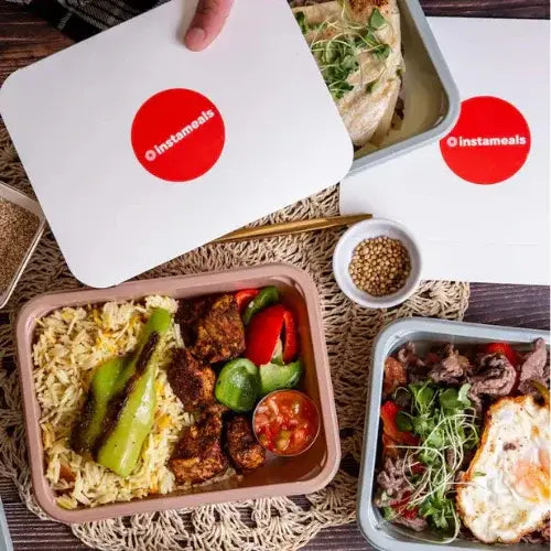 A flat lay of prepared meal containers from Instameals, with one being opened to reveal rice, grilled vegetables, and seasoned meat. Another tray includes beef slices, greens, and a sunny-side-up egg, while a third features a wrap with sprouts. The meals are neatly arranged on a woven placemat with a small bowl of seeds beside them.