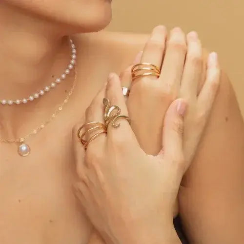 Close-up of a woman's hands and neckline adorned with gold statement rings and delicate pearl necklaces. The background is a warm beige, highlighting the elegant jewelry and soft skin tones.