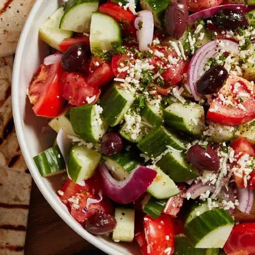 Greek salad with cucumbers, tomatoes, olives, and cheese in a white bowl.
