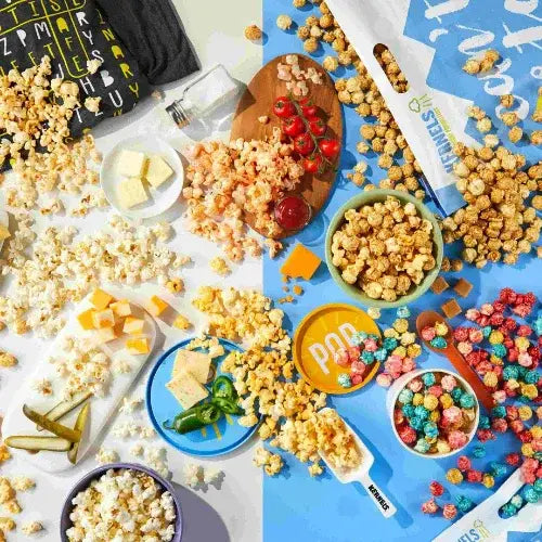 A colourful flat lay display of assorted popcorn varieties and snack ingredients, including buttered, caramel, cheddar, and rainbow popcorn. Surrounding items include cheese cubes, cherry tomatoes, jalapeños, crackers, and branded packaging on a bright split background of blue and white.