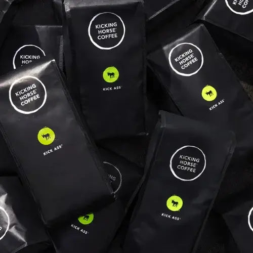 Multiple black bags of Kicking Horse Coffee arranged in a scattered flat lay. Each bag is labeled with the brand’s white circular logo and features a green circle with a black horse silhouette and the bold text 'Kick Ass®' beneath it.