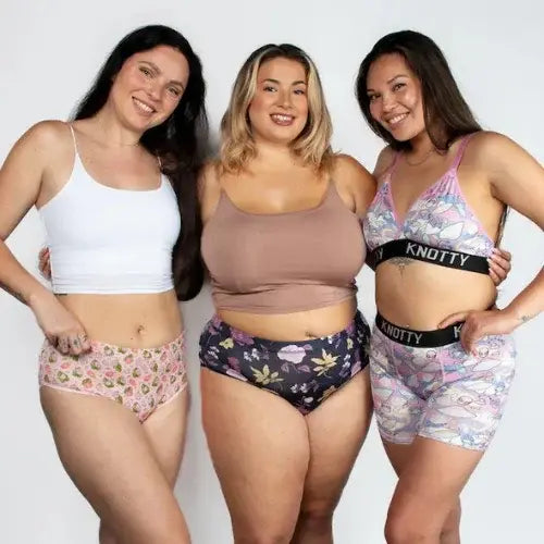 Three smiling women of diverse body types posing confidently in colourful, patterned underwear sets and crop tops. The underwear features fun prints including strawberries, florals, and unicorns, with the brand name 'KNOTTY' visible on the waistbands of two sets.