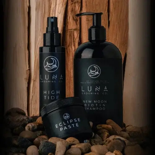 Three black bottles labeled 'High Tide', 'New Moon Biotin Shampoo', and 'Eclipse Paste' on a wooden surface with pebbles.