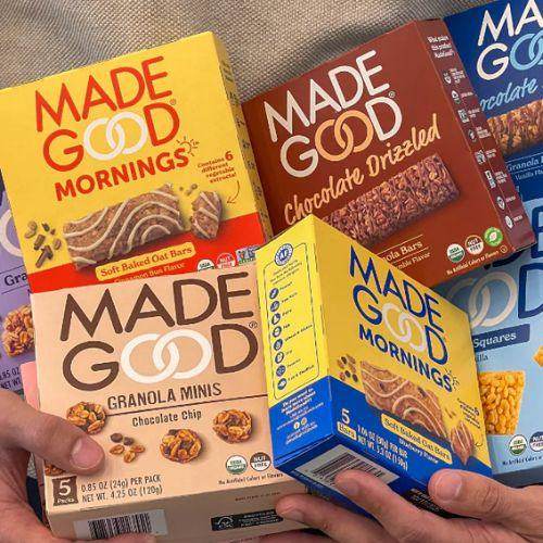 Hands holding various Made Good snack boxes including granola minis, chocolate drizzled bars, and soft baked oat bars in bright, colourful packaging