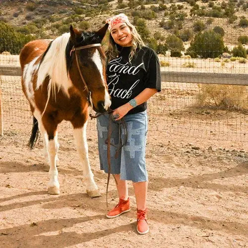 A smiling woman standing beside a brown and white horse in a sunny, desert-like setting. She wears a black 'Land Back' t-shirt, a red bandana, turquoise jewelry, a denim skirt with Indigenous-inspired designs, and red moccasin-style shoes