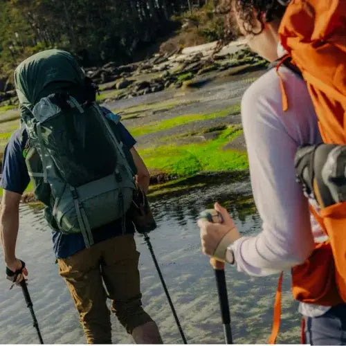 Two hikers with backpacks and walking sticks standing by a body of water.