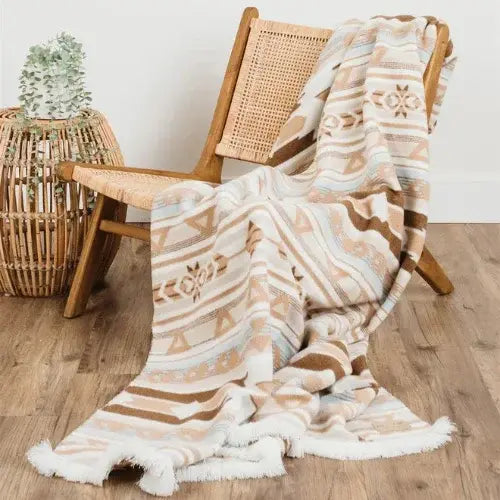Patterned blanket draped over a wooden chair with a woven side table and plant in the background.