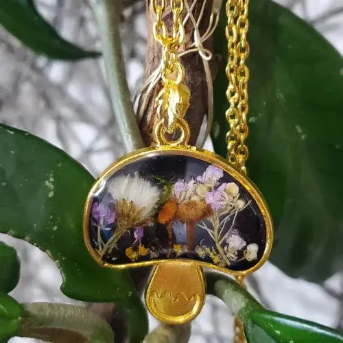 Gold necklace with a pendant featuring dried flowers and leaves on green leaves.