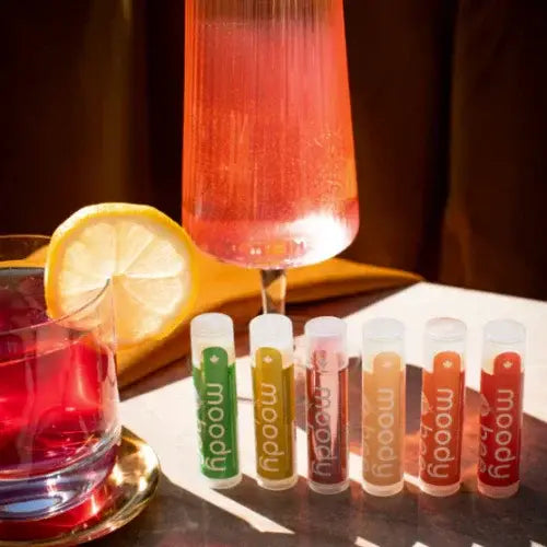 Set of colorful lip balms labeled 'moody' with a glass of red liquid and lemon slice on a wooden surface.
