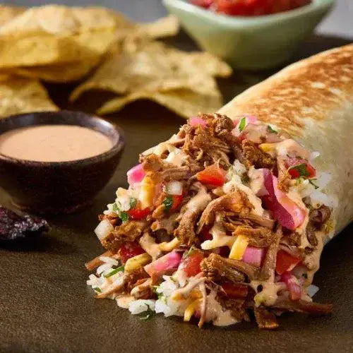 A grilled burrito overflowing with shredded beef, rice, diced tomatoes, onions, cheese, and creamy sauce, served on a dark plate with a side of tortilla chips, salsa, and dipping sauce in small bowls.
