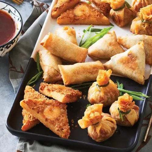 A platter of assorted crispy Asian appetizers, including spring rolls, samosas, fried wonton pouches, and shrimp toast triangles. Garnished with green onions and served with a bowl of dipping sauce, the appetizers are arranged on a two-toned black and white tray for an elegant presentation.