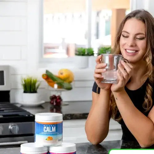 Woman holding a glass of water with a container labeled 'Natural Calm' on a kitchen counter.