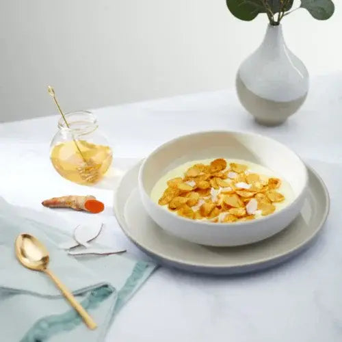 Bowl of cereal with yellow flakes on a table with a vase and spoon.
