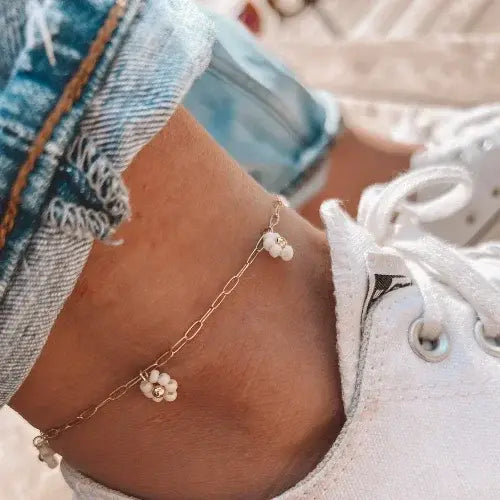 Close-up of a person's ankle wearing a gold anklet with floral charms, paired with white sneakers and denim shorts.