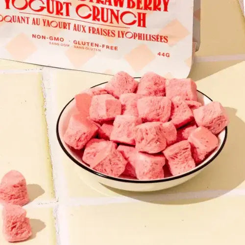 Pink freeze dried fruit pieces in a bowl with a package labelled 'Strawberry Crunch' on a yellow background