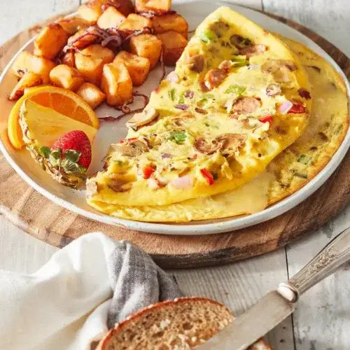 Vegetable omelette filled with mushrooms, peppers, and onions, served with home fries, a slice of orange, a strawberry, and a piece of whole grain toast on a rustic wooden board.