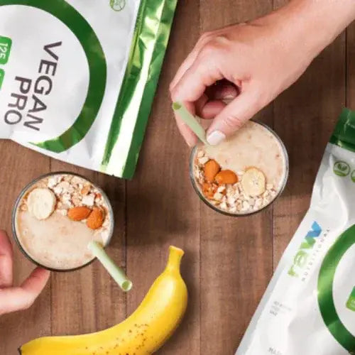 Two glasses of a vegan protein shake with a banana and packages of Vegan Pro on a wooden surface.