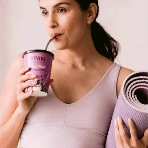 Woman drinking from a purple 'REVIVE' cup while holding a yoga mat.