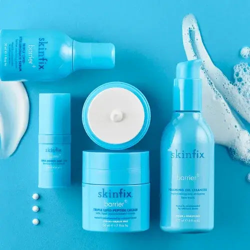 A flat lay of bright blue Skinfix skincare products from the Barrier+ line, including a foaming oil cleanser, triple lipid-peptide cream, and hydrating sprays. The products are surrounded by foam and cream textures on a matching blue background, creating a clean and refreshing aesthetic