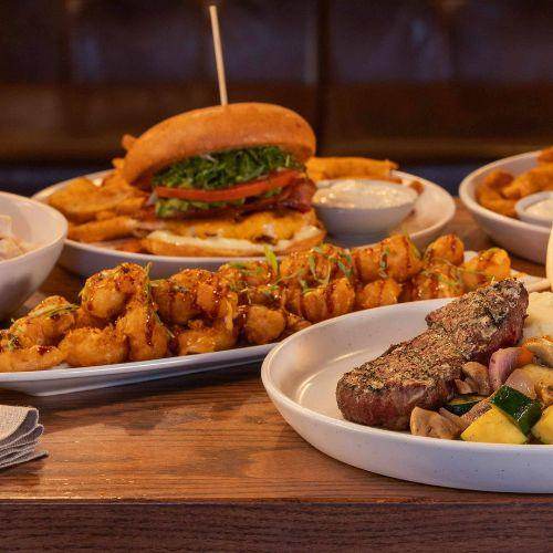 State & Main restaurant dishes including a gourmet burger, steak with vegetables, and crispy appetizers on a wooden table